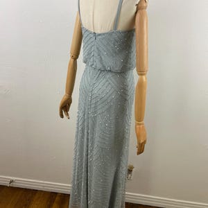K&ouml;nnte beinhalten: Ein hellblaues, bodenlanges Kleid mit Perlenmuster. Das Kleid hat d&uuml;nne Tr&auml;ger und ein eng anliegendes Oberteil mit flie&szlig;endem Rock. Die Perlen erzeugen ein subtiles Muster auf dem Stoff. Das Kleid wird auf einer Schaufensterpuppe pr&auml;sentiert.