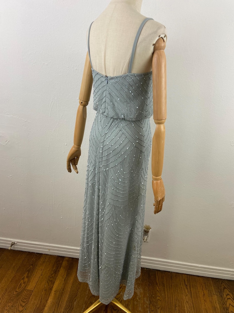 K&ouml;nnte beinhalten: Ein hellblaues, bodenlanges Kleid mit Perlenmuster. Das Kleid hat d&uuml;nne Tr&auml;ger und ein eng anliegendes Oberteil mit flie&szlig;endem Rock. Die Perlen erzeugen ein subtiles Muster auf dem Stoff. Das Kleid wird auf einer Schaufensterpuppe pr&auml;sentiert.