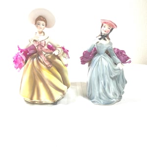May include: Two porcelain figurines of women in vintage dresses, each holding a bouquet of pink roses. One wears a yellow dress and a wide-brimmed hat, the other a blue dress and a small hat.