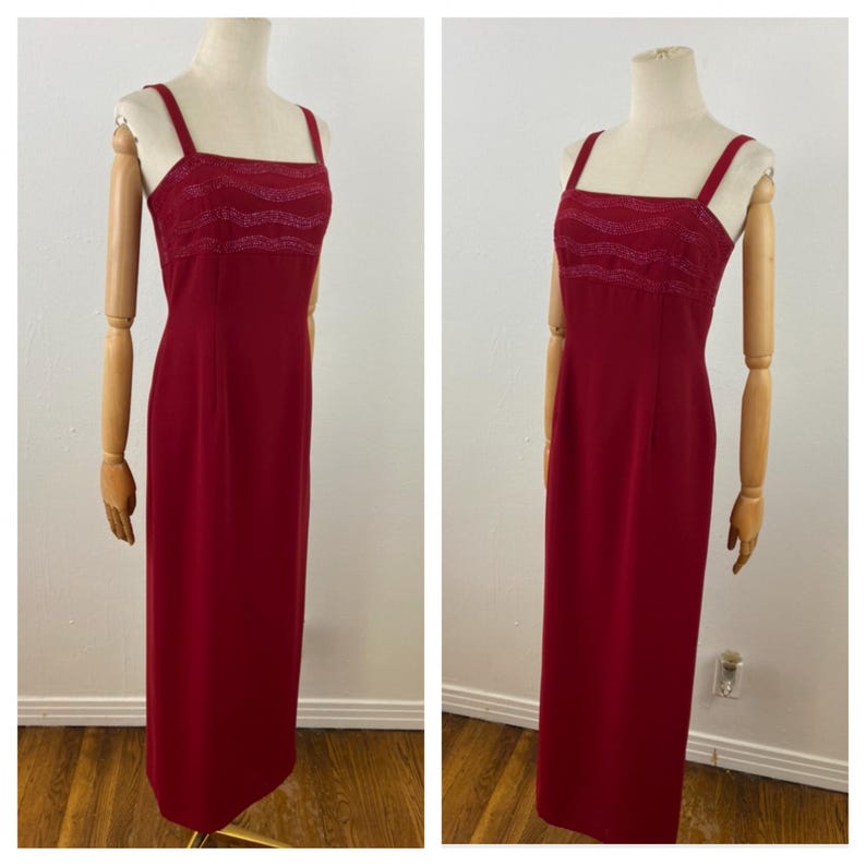 Op de afbeelding: Een lange, rode avondjurk met een vierkante halslijn en dunne bandjes. De bodice heeft een gestructureerd, golfachtig ontwerp. De jurk wordt op een mannequin getoond en toont zijn vloerlange silhouet. De jurk heeft een effen, dieprode kleur.