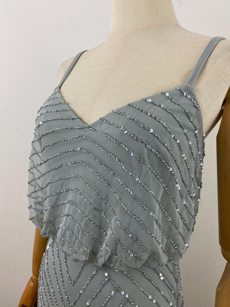 K&ouml;nnte beinhalten: Hellblaues Kleid mit V-Ausschnitt und Spaghettitr&auml;gern. Das Kleid ist mit Reihen silberner Pailletten in einem Zickzackmuster verziert. Das Oberteil hat ein mehrschichtiges Design, das dem eleganten Abendkleid Tiefe verleiht.