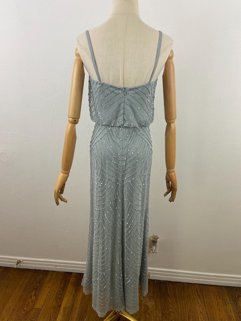 K&ouml;nnte beinhalten: Ein hellblaues Abendkleid mit Perlenbesatz. Das Kleid hat d&uuml;nne Spaghettitr&auml;ger und ein eng anliegendes Oberteil, das in einen bodenlangen Rock &uuml;bergeht. Die R&uuml;ckseite des Kleides ist sichtbar und zeigt den Rei&szlig;verschluss und die aufwendigen Perlenarbeiten.