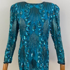 May include: Teal-colored long-sleeved top heavily embellished with sequins and beads. The intricate design features a pattern of large, iridescent sequins and silver beading, creating a shimmering effect. The top is displayed on a mannequin, showcasing its detailed craftsmanship.
