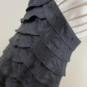 May include: A close-up of a black, layered fabric. The fabric appears to be a dress or top, with multiple horizontal layers creating a ruffled effect. The texture of the fabric is slightly shiny, and the layers cascade down the garment.