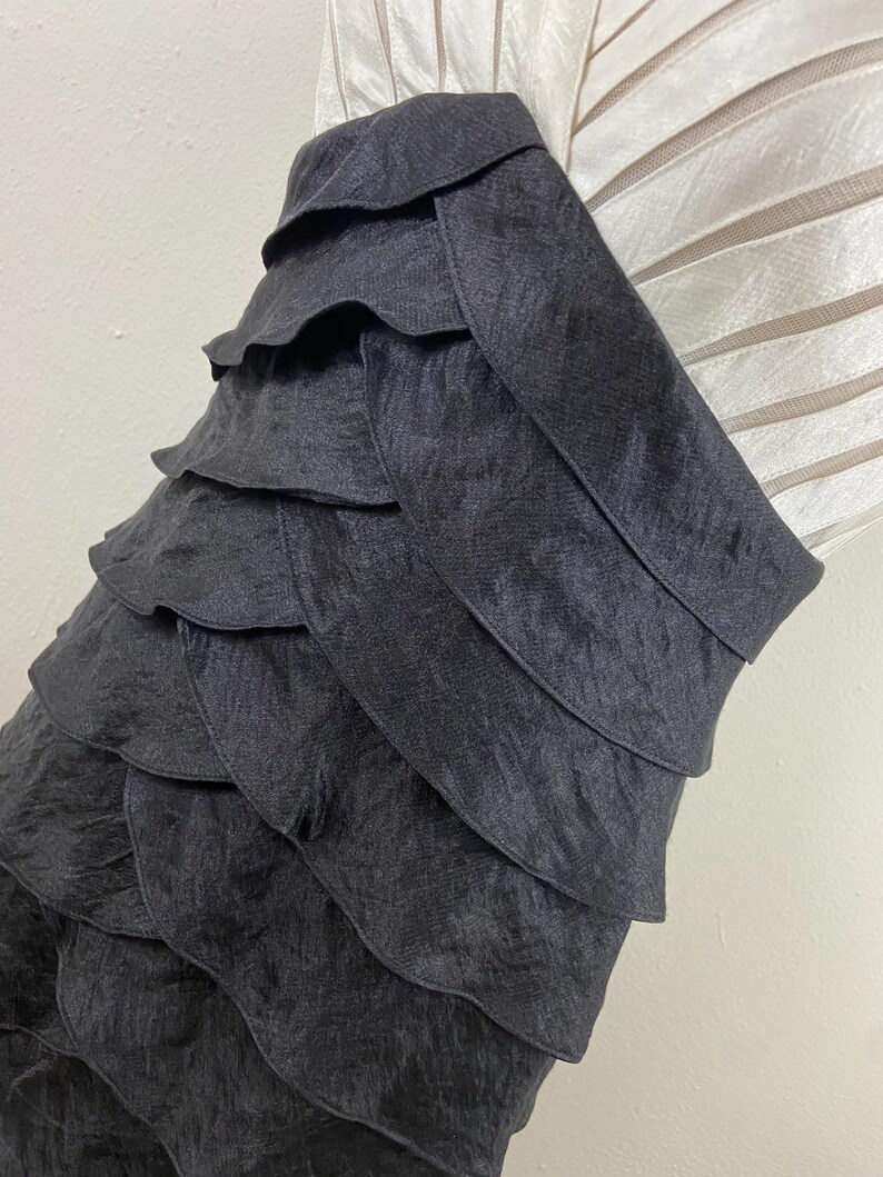 May include: A close-up of a black, layered fabric. The fabric appears to be a dress or top, with multiple horizontal layers creating a ruffled effect. The texture of the fabric is slightly shiny, and the layers cascade down the garment.