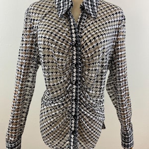 May include: A long-sleeved button-down shirt with a houndstooth pattern in black, white, and tan. The shirt has a classic collar and ruched detailing down the front. The cuffs are buttoned.