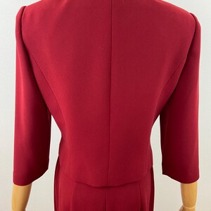 Op de afbeelding: Een rode tweedelige outfit, bestaande uit een blazer met driekwart mouwen en een bijpassende rok. De outfit wordt getoond op een mannequin, met de achterkant van de jas en rok. De stof lijkt een effen, glad materiaal te zijn.