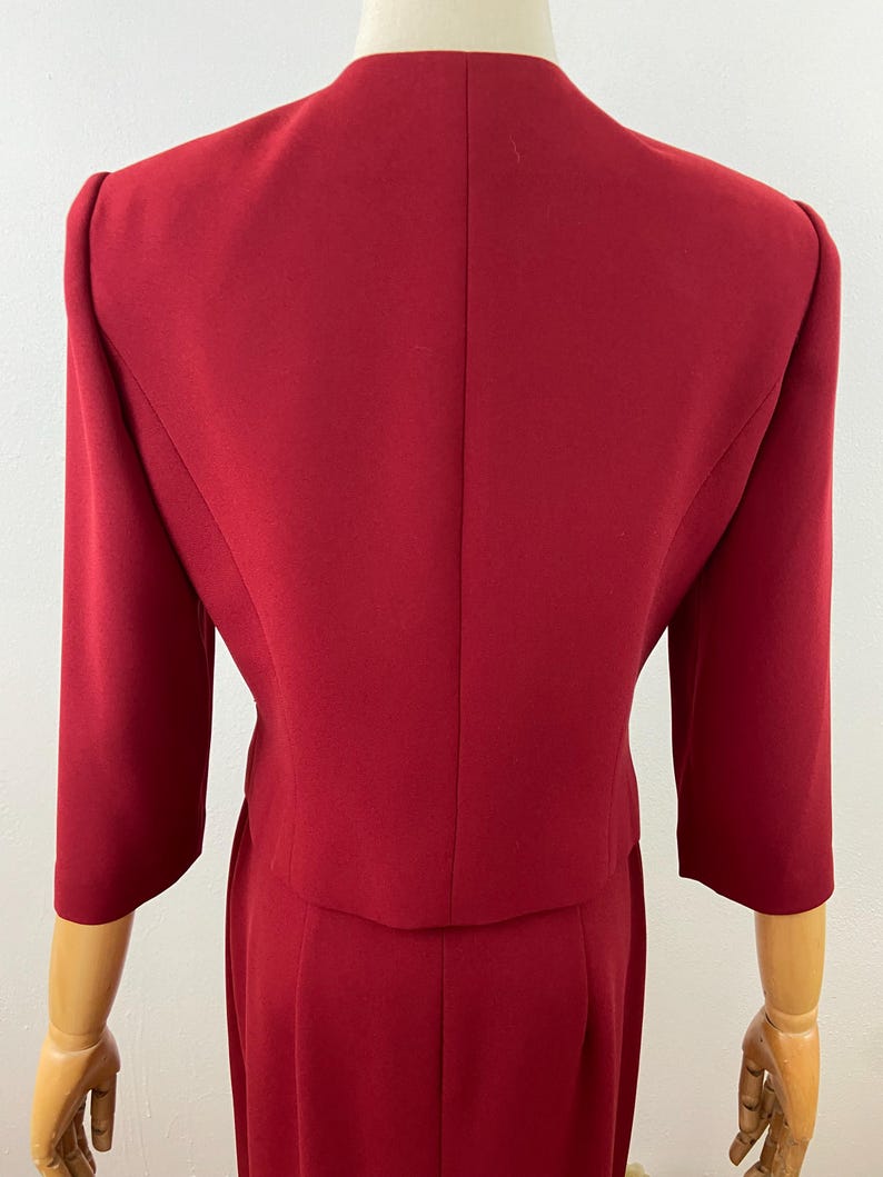 Op de afbeelding: Een rode tweedelige outfit, bestaande uit een blazer met driekwart mouwen en een bijpassende rok. De outfit wordt getoond op een mannequin, met de achterkant van de jas en rok. De stof lijkt een effen, glad materiaal te zijn.