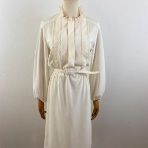 Peut inclure: Robe vintage crème avec manches longues et jupe mi-longue. La robe présente un col haut à volants, des plis verticaux et des détails en dentelle sur le corsage. Une ceinture assortie cintre la taille, présentée sur un mannequin.