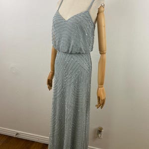 K&ouml;nnte beinhalten: Ein hellblaues, bodenlanges Kleid mit V-Ausschnitt und Spaghettitr&auml;gern. Das Kleid ist mit silbernen Pailletten in einem geometrischen Muster verziert. Das Kleid wird auf einer Schaufensterpuppe pr&auml;sentiert.