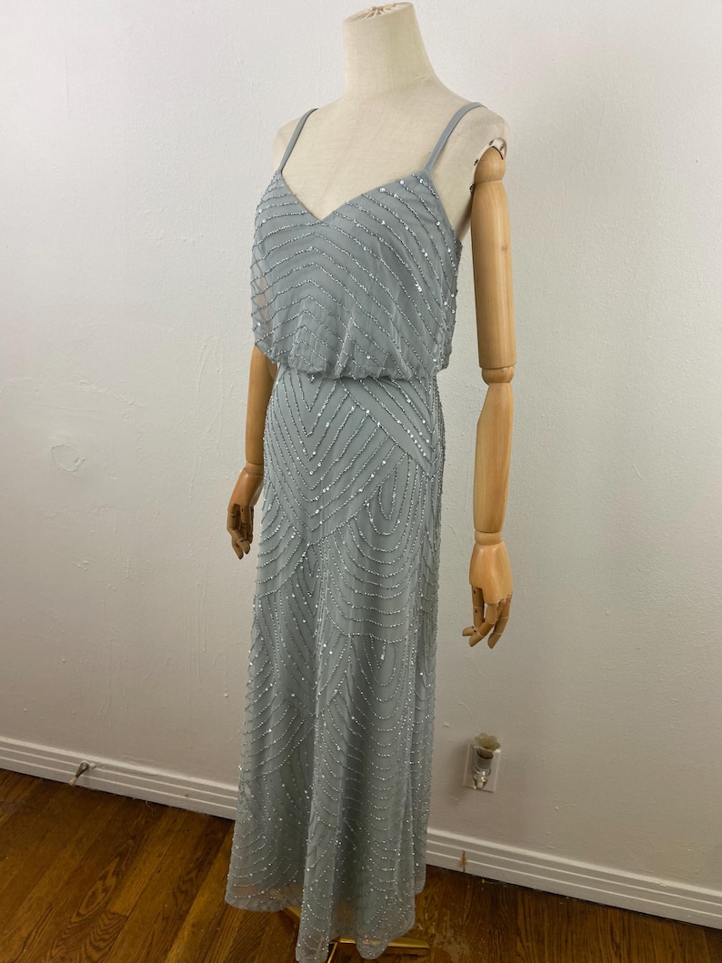 K&ouml;nnte beinhalten: Ein hellblaues, bodenlanges Kleid mit V-Ausschnitt und Spaghettitr&auml;gern. Das Kleid ist mit silbernen Pailletten in einem geometrischen Muster verziert. Das Kleid wird auf einer Schaufensterpuppe pr&auml;sentiert.