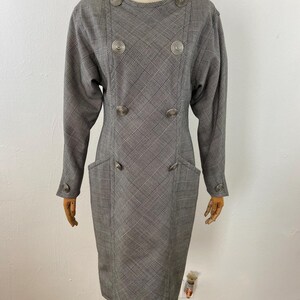 May include: A vintage gray and white plaid dress with long sleeves and a knee-length hem. The dress features large, decorative buttons and two front pockets. The dress is displayed on a mannequin.