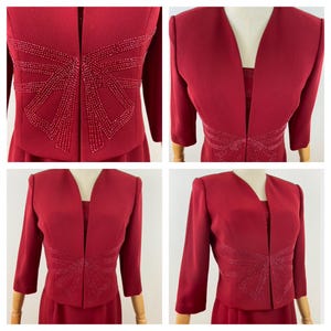 Op de afbeelding: Een rode tweedelige outfit met een jasje en een jurk. Het jasje heeft driekwart mouwen en een V-hals. Zowel het jasje als de jurk zijn versierd met een kralenpatroon. De outfit wordt gepresenteerd op een mannequin.