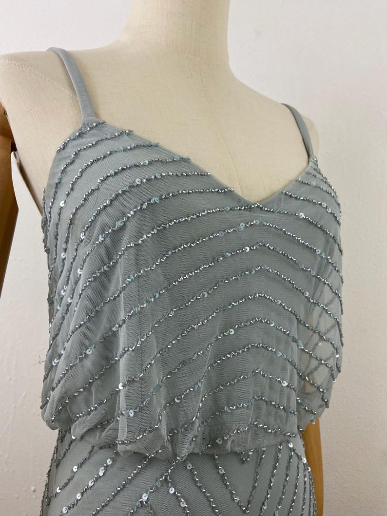 K&ouml;nnte beinhalten: Hellblaues Kleid mit V-Ausschnitt und Spaghettitr&auml;gern. Das Kleid ist mit Reihen aus silbernen Pailletten verziert, die einen schimmernden Effekt erzeugen. Das Kleid wird auf einer Schaufensterpuppe pr&auml;sentiert.