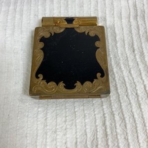 May include: A vintage compact with a rectangular shape and a hinged lid. The compact features a black center panel framed by ornate gold-colored filigree. A small, cylindrical component is attached to the top.