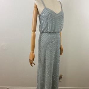 K&ouml;nnte beinhalten: Ein hellblaues, bodenlanges Kleid mit V-Ausschnitt und Spaghettitr&auml;gern. Das Kleid ist mit silbernen Perlen in einem linearen und geometrischen Muster verziert. Das Kleid wird auf einer Schaufensterpuppe pr&auml;sentiert.