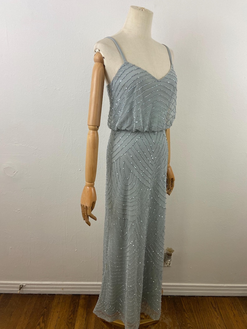 K&ouml;nnte beinhalten: Ein hellblaues, bodenlanges Kleid mit V-Ausschnitt und Spaghettitr&auml;gern. Das Kleid ist mit silbernen Perlen in einem linearen und geometrischen Muster verziert. Das Kleid wird auf einer Schaufensterpuppe pr&auml;sentiert.