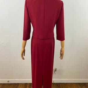 Op de afbeelding: Een tweedelig rood broekpak. De jas heeft driekwart mouwen en een ronde kraag. De rok is een rechte, vloerlange stijl met een split aan de achterkant. Het pak wordt op een mannequin gepresenteerd.