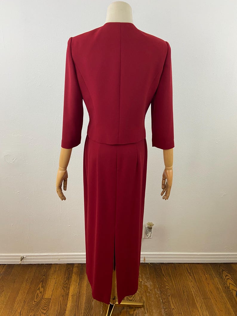 Op de afbeelding: Een tweedelig rood broekpak. De jas heeft driekwart mouwen en een ronde kraag. De rok is een rechte, vloerlange stijl met een split aan de achterkant. Het pak wordt op een mannequin gepresenteerd.