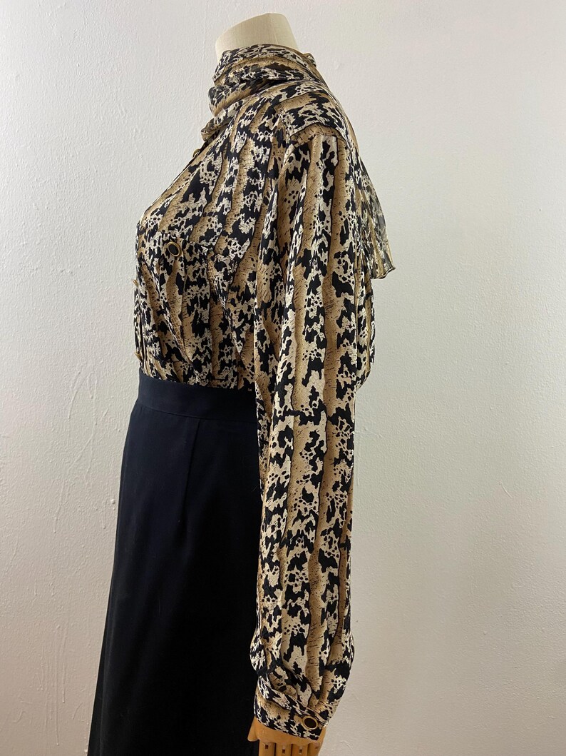 Peut inclure: Ensemble vintage compos&eacute; d'un chemisier et d'une jupe. Le chemisier pr&eacute;sente un col montant et des manches longues avec un imprim&eacute; animalier beige, noir et cr&egrave;me. La jupe est noire unie, de longueur genou. L'ensemble est pr&eacute;sent&eacute; sur un mannequin.