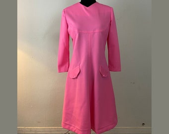May include: A pink vintage dress with long sleeves and two pockets. The dress has a simple design and a slightly flared skirt.