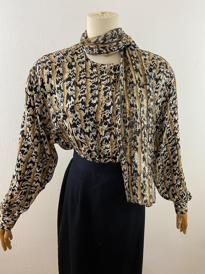 Peut inclure: Ensemble vintage compos&eacute; d'un chemisier et d'une jupe. Le chemisier pr&eacute;sente un imprim&eacute; animalier marron, noir et beige avec des manches longues et une &eacute;charpe assortie. La jupe est noire unie, taille haute. Le chemisier a des boutons dor&eacute;s.