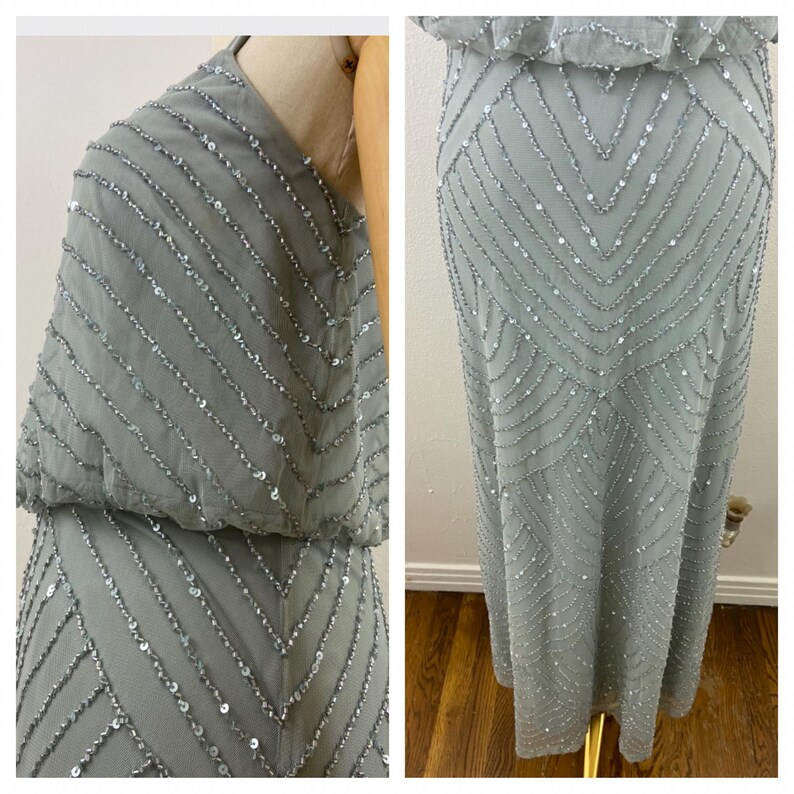 K&ouml;nnte beinhalten: Ein hellgraues Abendkleid mit aufwendigen Pailletten- und Perlenverzierungen. Das Kleid hat ein eng anliegendes Oberteil und einen flie&szlig;enden Rock mit einem Muster aus diagonalen und Zickzacklinien. Das Kleid wird auf einer Schaufensterpuppe pr&auml;sentiert.