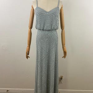 K&ouml;nnte beinhalten: Ein hellblaues, bodenlanges Kleid mit V-Ausschnitt und Spaghettitr&auml;gern. Das Kleid ist mit silbernen Perlen verziert, die in horizontalen und Zickzackmustern angeordnet sind. Das Oberteil hat eine taillierte Taille, und der Rock f&auml;llt bis zum Boden.