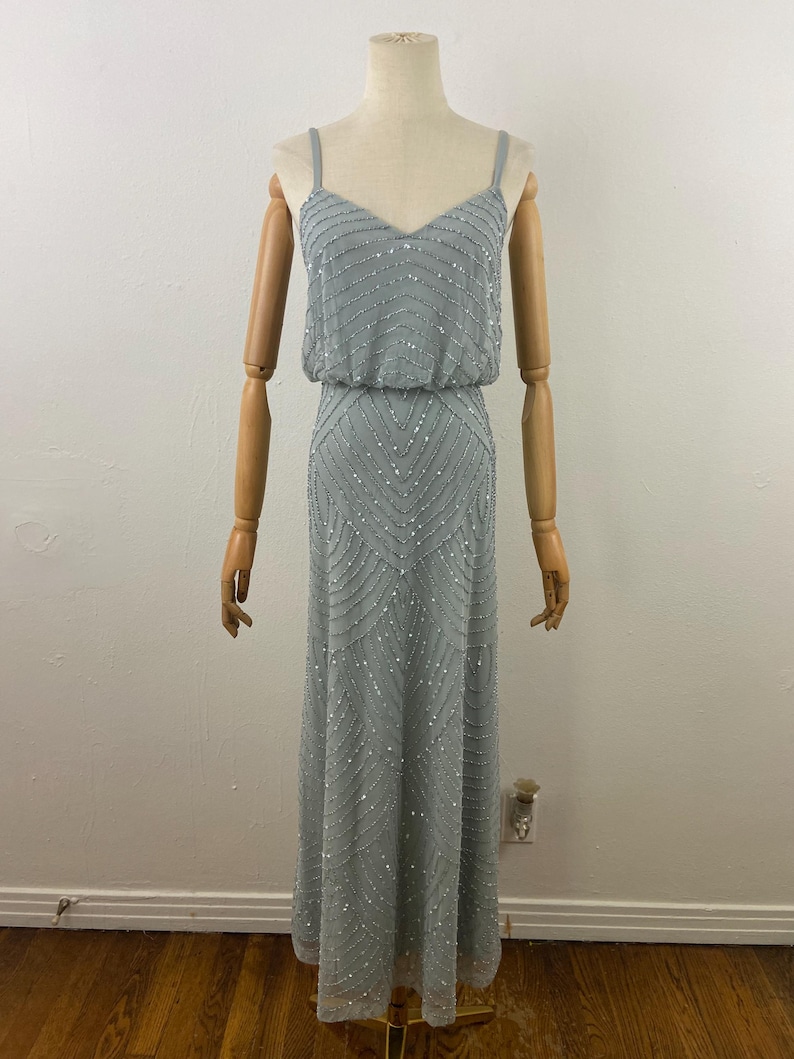 K&ouml;nnte beinhalten: Ein hellblaues, bodenlanges Kleid mit V-Ausschnitt und Spaghettitr&auml;gern. Das Kleid ist mit silbernen Perlen verziert, die in horizontalen und Zickzackmustern angeordnet sind. Das Oberteil hat eine taillierte Taille, und der Rock f&auml;llt bis zum Boden.