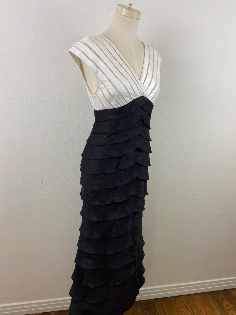 May include: A formal, floor-length dress with a layered black skirt and a white bodice. The bodice features a V-neck and horizontal striped detailing. The dress is displayed on a mannequin.