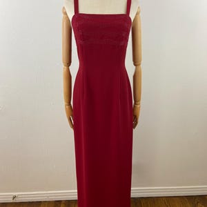 Op de afbeelding: Een lange, rode avondjurk met een vierkante halslijn en dunne bandjes. De bodice heeft een kralenontwerp. De jurk wordt getoond op een mannequin met houten armen en benen, tegen een witte muur en houten vloer.
