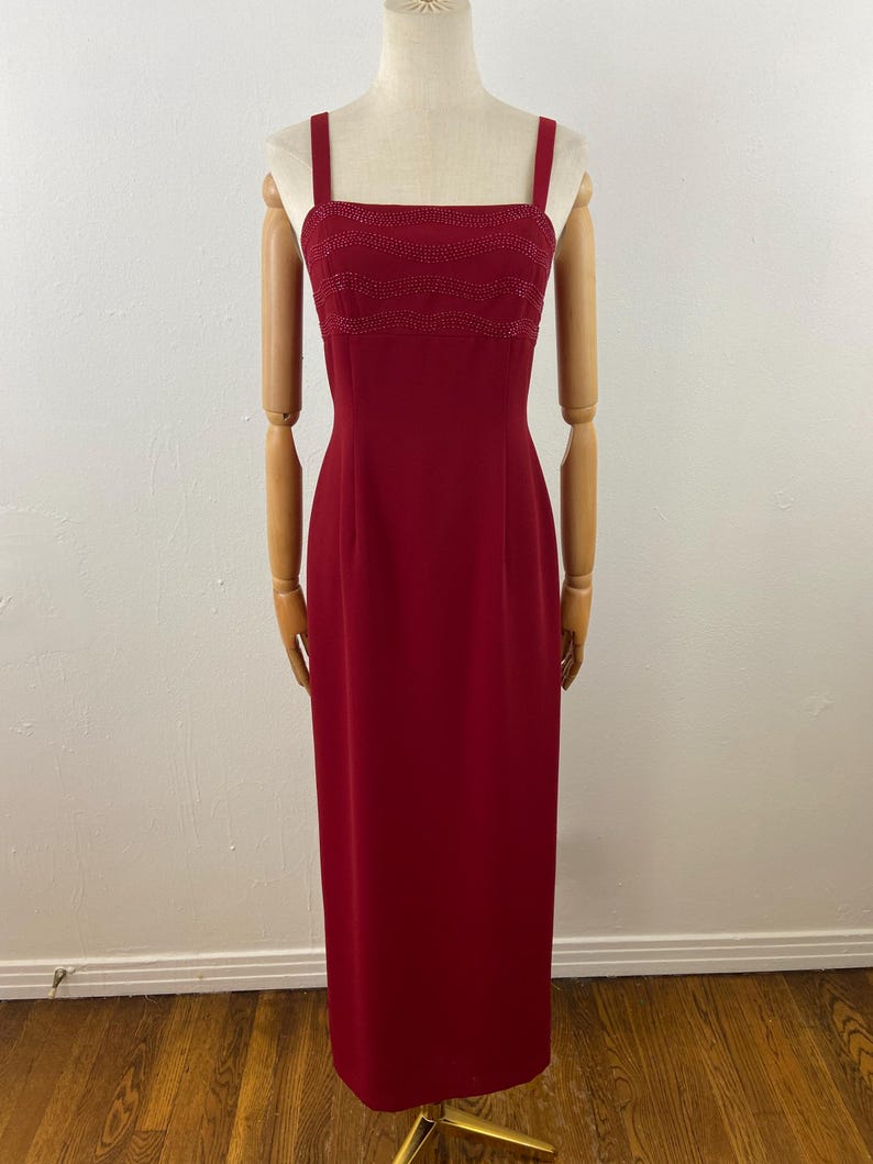 Op de afbeelding: Een lange, rode avondjurk met een vierkante halslijn en dunne bandjes. De bodice heeft een kralenontwerp. De jurk wordt getoond op een mannequin met houten armen en benen, tegen een witte muur en houten vloer.