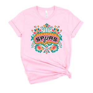 May include: A light pink t-shirt featuring a colorful floral design with the words "San Antonio Spurs" in a rectangular frame. The design includes orange, teal, and white accents. The shirt has rolled-up sleeves.
