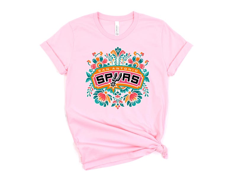 May include: A light pink t-shirt featuring a colorful floral design with the words "San Antonio Spurs" in a rectangular frame. The design includes orange, teal, and white accents. The shirt has rolled-up sleeves.