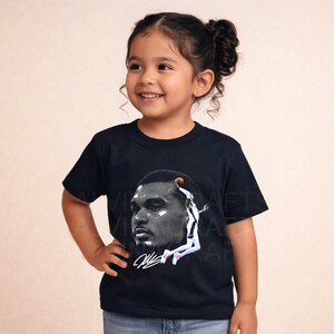May include: A navy blue t-shirt featuring a black and white image of a basketball player. The image includes a signature and a basketball. The t-shirt is being worn by a young child.