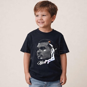 May include: A black t-shirt featuring a grayscale image of a basketball player in action. The graphic includes a portrait of the player and a depiction of him jumping to shoot a basketball. The shirt is worn by a young child.