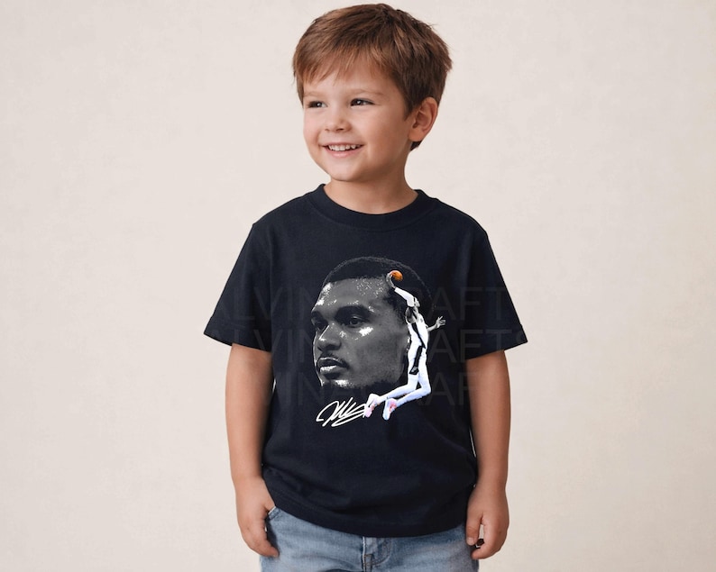 May include: A black t-shirt featuring a grayscale image of a basketball player in action. The graphic includes a portrait of the player and a depiction of him jumping to shoot a basketball. The shirt is worn by a young child.