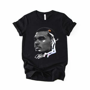 May include: Black t-shirt featuring a grayscale image of a basketball player's face and a leaping figure with a basketball. The shirt also includes a white signature. The design is centered on the front of the shirt.