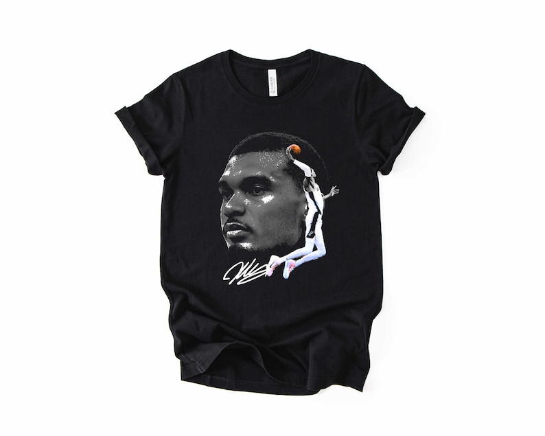 May include: Black t-shirt featuring a grayscale image of a basketball player's face and a leaping figure with a basketball. The shirt also includes a white signature. The design is centered on the front of the shirt.