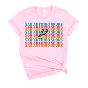 Puede incluir: Camiseta rosa claro con la inscripci&oacute;n "SAN ANTONIO SPURS" repetida en un patr&oacute;n de color turquesa, naranja y blanco. El logotipo de los Spurs de San Antonio est&aacute; en el centro. La camiseta es de tejido suave.