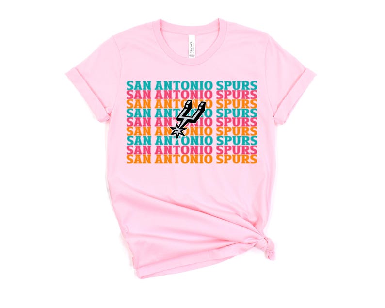 Puede incluir: Camiseta rosa claro con la inscripci&oacute;n "SAN ANTONIO SPURS" repetida en un patr&oacute;n de color turquesa, naranja y blanco. El logotipo de los Spurs de San Antonio est&aacute; en el centro. La camiseta es de tejido suave.