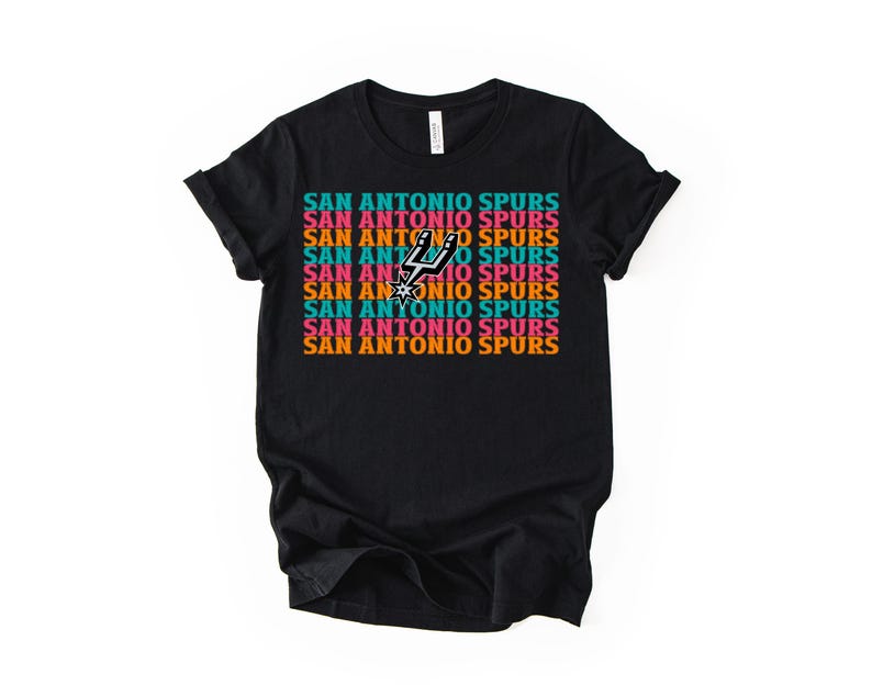 May include: Black t-shirt featuring the text "SAN ANTONIO SPURS" in a repeating pattern of teal, orange, pink, and white. The Spurs logo is centered on the shirt.