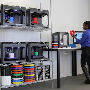 May include: A woman stands in a room with several 3D printers. The printers are on a metal shelf and are black with a clear plastic build area. The woman is holding a red 3D printed object in her hand. There are spools of filament in various colors on the bottom shelf.
