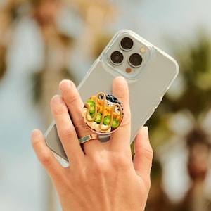 May include: A circular phone grip featuring a miniature food bowl design with colorful toppings including green slices, yellow strands, and dark blue berries, drizzled with a tan sauce, attached to the back of a clear smartphone case.