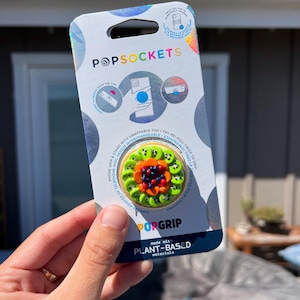 May include: A PopSockets phone grip with a colorful design featuring kiwi slices, orange petals, and dark berries. The packaging includes the PopSockets logo and text indicating it's made with plant-based materials.