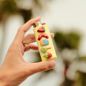 Pu&ograve; includere: Un accendino giallo decorato con ornamenti colorati in rilievo a tema cibo. Le decorazioni includono una fetta di anguria, una salsiccia, un gelato, un lecca-lecca e una fragola. L'accendino &egrave; tenuto in mano.