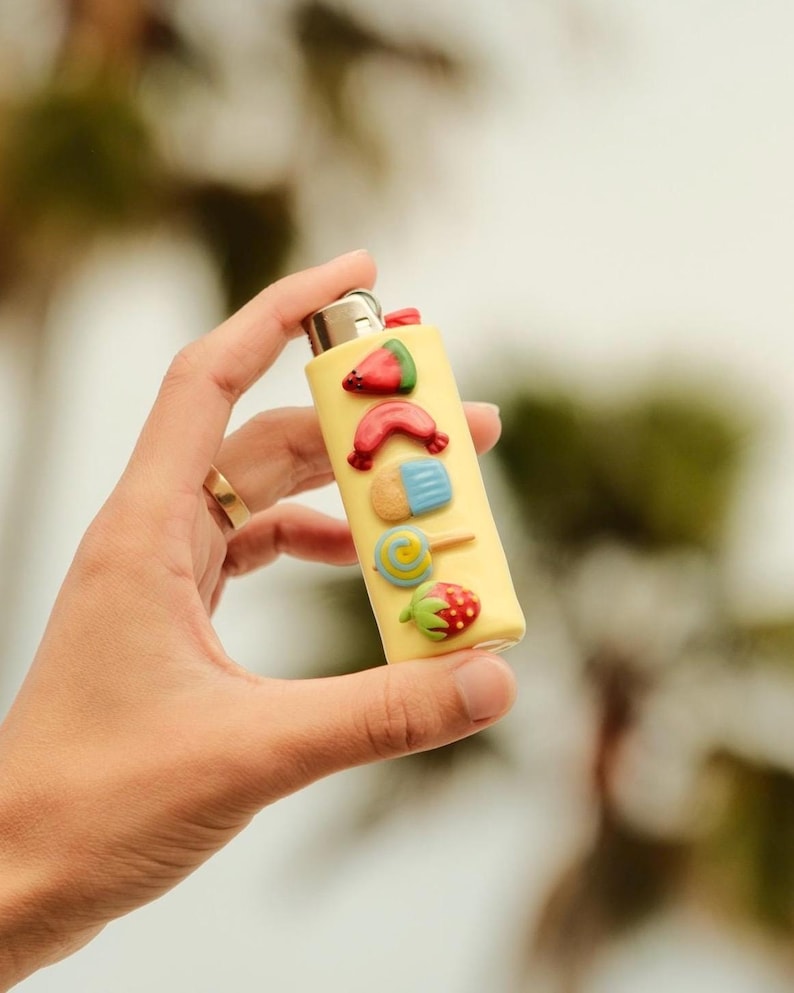 Pu&ograve; includere: Un accendino giallo decorato con ornamenti colorati in rilievo a tema cibo. Le decorazioni includono una fetta di anguria, una salsiccia, un gelato, un lecca-lecca e una fragola. L'accendino &egrave; tenuto in mano.