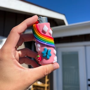 Pu&ograve; includere: Un accendino rosa con un arcobaleno, forme di diamante bianco e un design a quadrifoglio blu. &Egrave; visibile anche una fragola rossa. L'accendino &egrave; tenuto contro un cielo blu brillante.