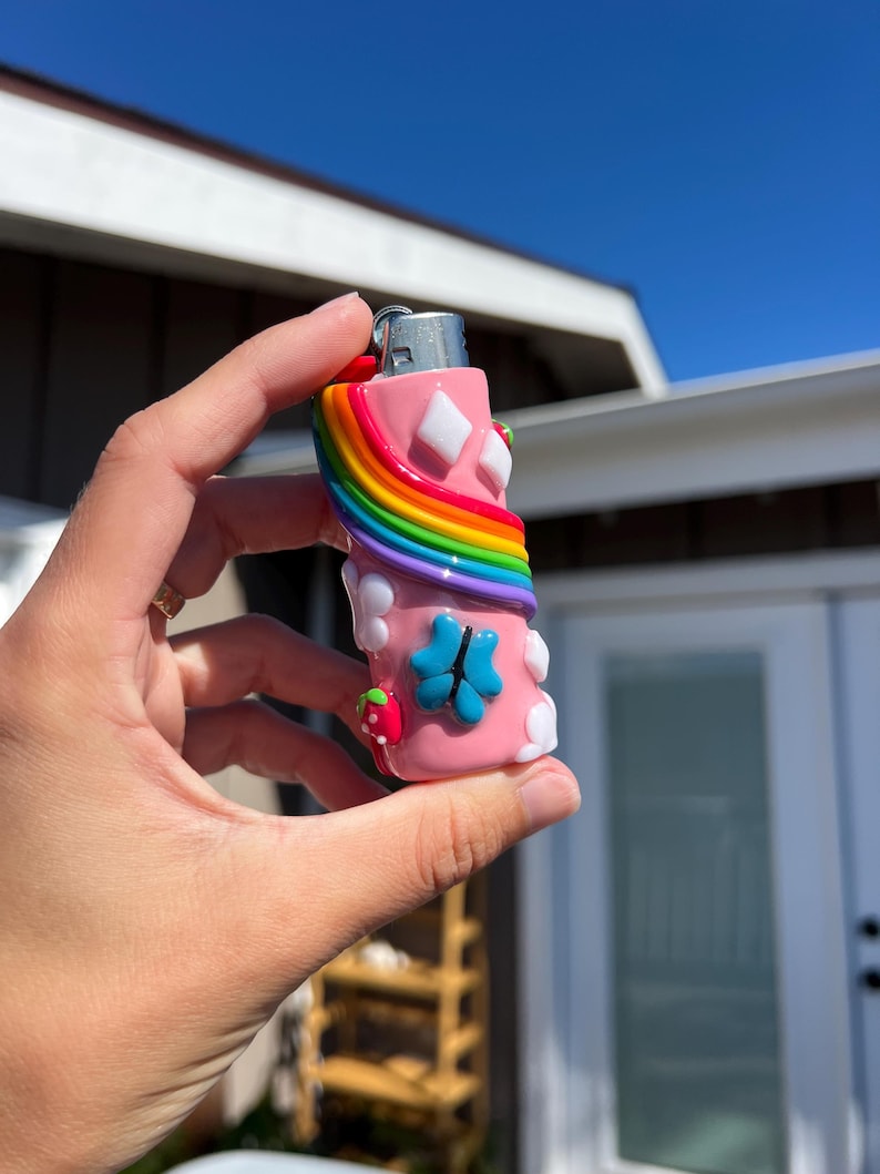 Pu&ograve; includere: Un accendino rosa con un arcobaleno, forme di diamante bianco e un design a quadrifoglio blu. &Egrave; visibile anche una fragola rossa. L'accendino &egrave; tenuto contro un cielo blu brillante.