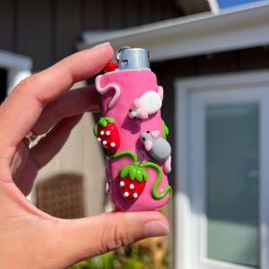 Pu&ograve; includere: Un accendino rosa decorato con fragole e piccoli topi bianchi e grigi. Le fragole hanno corpi rossi con semi bianchi e steli verdi. I topi sono in varie pose, aggiungendo un tocco stravagante all'accendino.